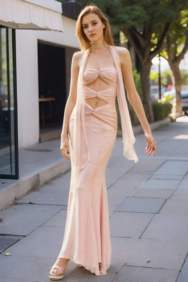 Woman wearing a pale pink halter neck maxi dress with cut-out details and a mermaid hemline, standing on a sidewalk. Main image