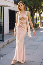 Woman wearing a pale pink halter neck maxi dress with cut-out details and a mermaid hemline, standing on a sidewalk.