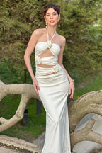 Woman wearing a white satin halter maxi dress with cut-out detailing and floral embellishments, posing on a tree branch