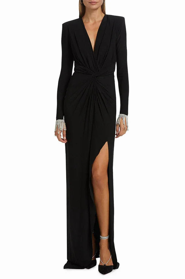Woman wearing a long-sleeved black jersey knit gown with a plunging V-neckline, ruched bodice, thigh-high slit, and embellished cuffs.