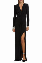 Woman wearing a long-sleeved black jersey knit gown with a plunging V-neckline, ruched bodice, thigh-high slit, and embellished cuffs.
