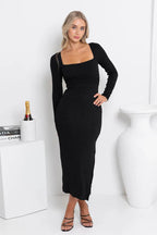 Woman wearing a black long-sleeved midi dress with a square neckline and side slit, posing with hand on hip