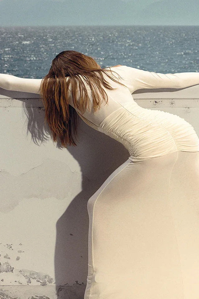 Woman in a cream sheer mesh gown leaning against a wall by the ocean.