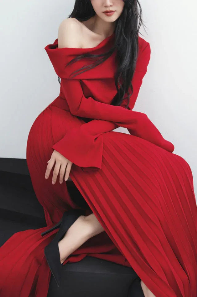Woman in a red off-the-shoulder dress with a pleated skirt and high slit, seated on a dark platform.