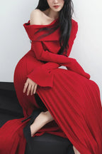 Woman in a red off-the-shoulder dress with a pleated skirt and high slit, seated on a dark platform.