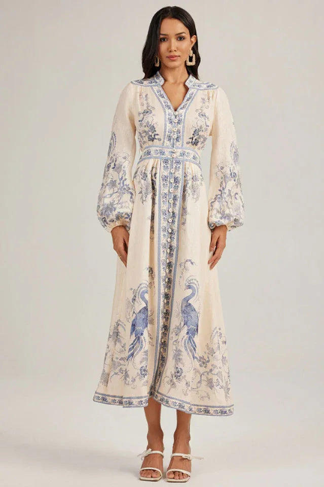 Full-length view of a woman in a long, cream-colored maxi dress with blue peacock motifs and a collared neckline. Secondary image