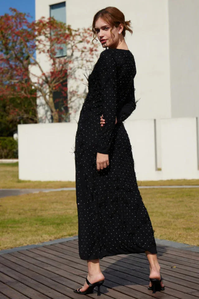 Back view of a woman in a black sparkly maxi dress with a round neckline and long sleeves.