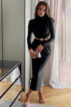 Woman wearing a long-sleeved black satin dress with a cut-out waist and embellished detail, holding a clutch purse and wearing high heels.