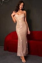 Woman in a champagne sequined formal gown, touching her chest, with spaghetti straps and a cowl neck.