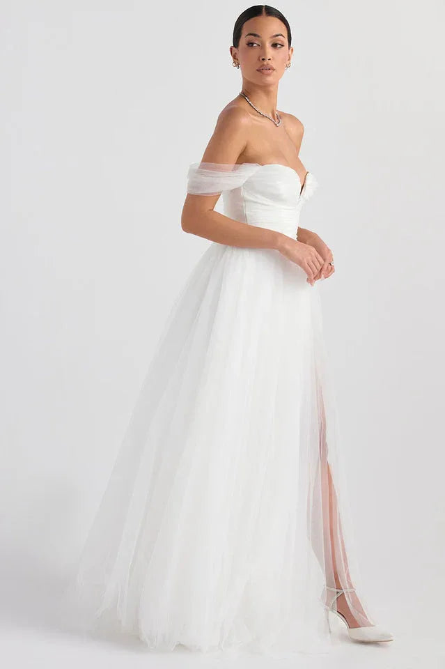 Model in a white off-the-shoulder wedding dress with a high slit, standing against a white background.