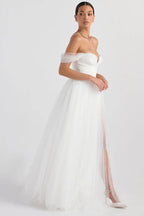 Model in a white off-the-shoulder wedding dress with a high slit, standing against a white background.