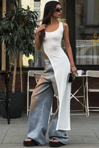 Woman wearing a white sleeveless maxi dress with a high slit over light blue wide-leg jeans and black platform sandals outdoors