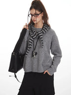 Woman wearing a grey knit sweater with a striped scarf, black cargo pants, and a black crossbody bag, holding her scarf.