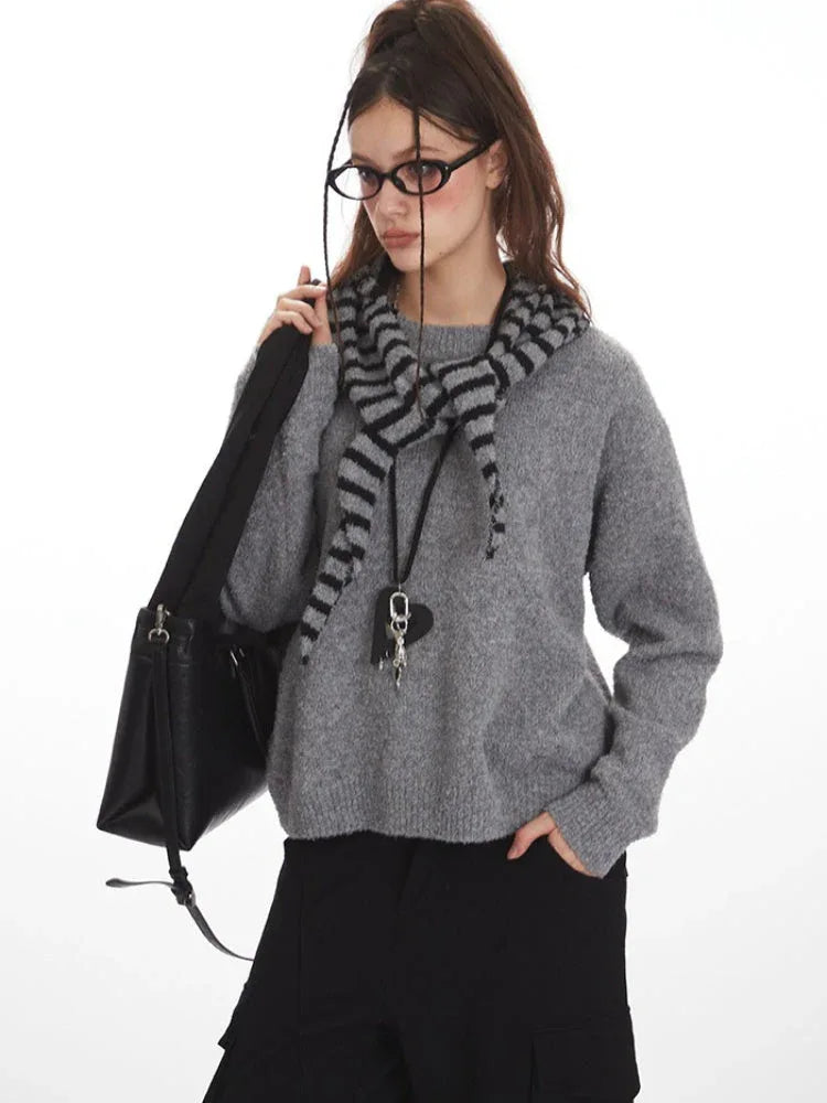 Woman wearing a grey knit sweater with a striped scarf, black cargo pants, and a black crossbody bag, holding her scarf.