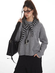 Woman wearing a grey knit sweater with a striped scarf, black cargo pants, and a black crossbody bag, holding her scarf.