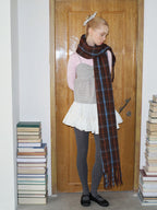 A young woman with blonde hair pulled back stands in front of a wooden door, wearing a pink long-sleeved shirt, a grey and white patterned top, a white skirt, grey polka dot tights, black shoes, and a long, plaid scarf, looking down.