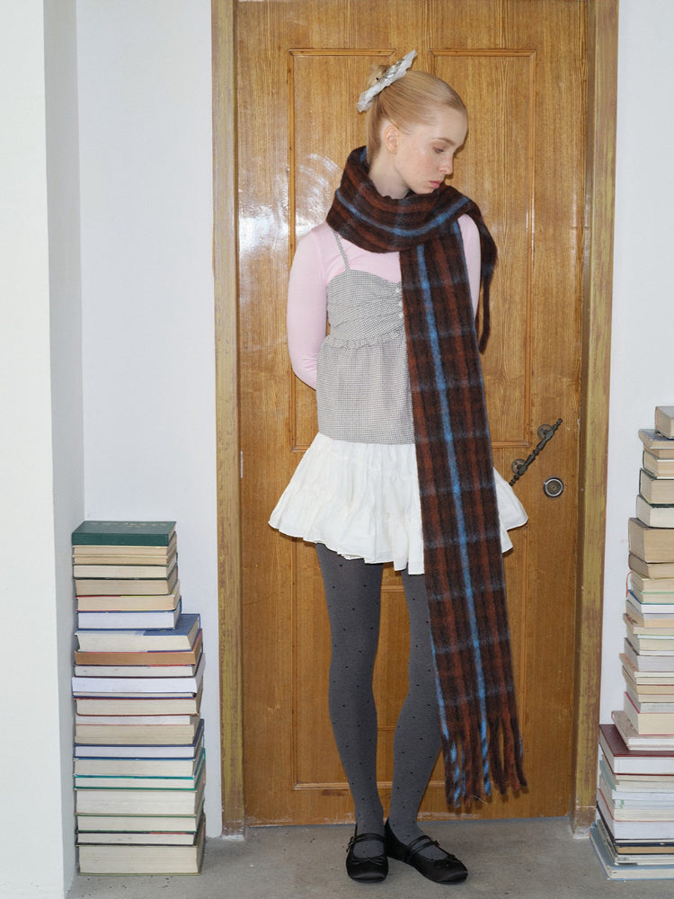 A young woman with blonde hair pulled back stands in front of a wooden door, wearing a pink long-sleeved shirt, a grey and white patterned top, a white skirt, grey polka dot tights, black shoes, and a long, plaid scarf, looking down.