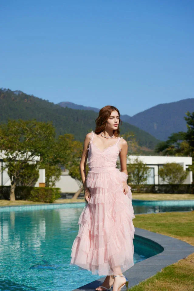 Woman in a light pink tiered tulle dress standing on the edge of a pool with mountains in the background.