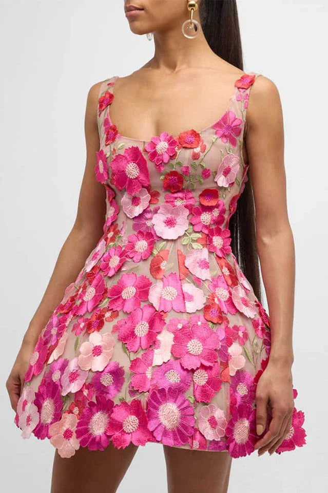 A woman wears a short, sleeveless dress adorned with vibrant pink and red floral appliqués over a sheer beige base. The dress features a scoop neckline and a flaring skirt.