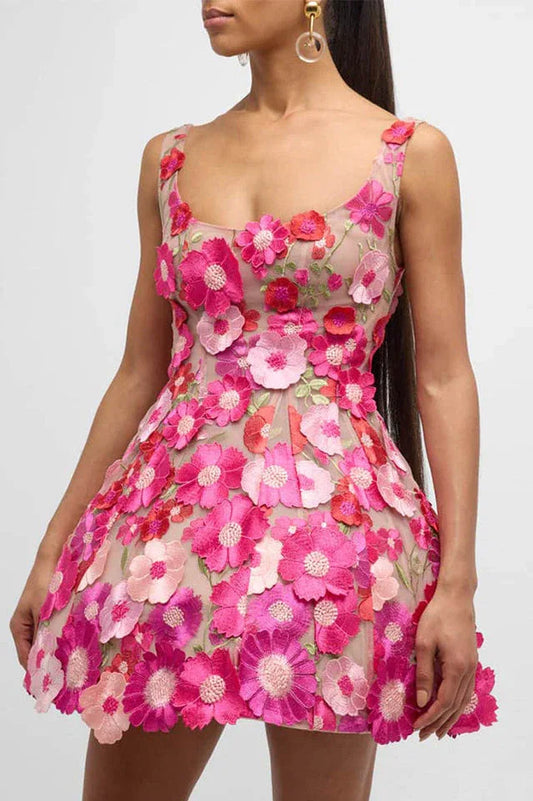A woman wears a short, sleeveless dress adorned with vibrant pink and red floral appliqués over a sheer beige base. The dress features a scoop neckline and a flaring skirt.