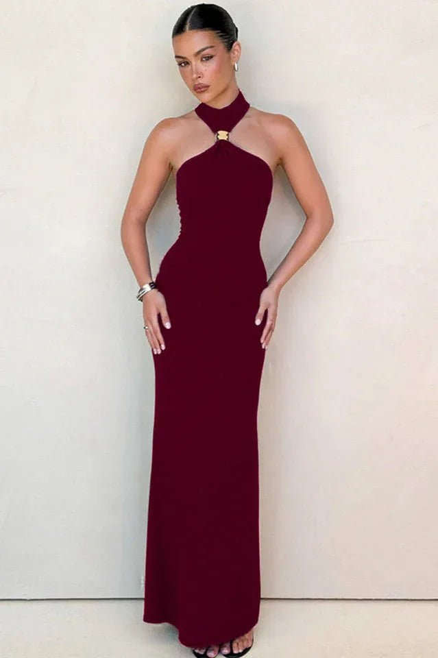 Woman wearing a burgundy sleeveless halter maxi dress with a gold-tone hardware detail at the neckline, standing against a neutral wall. Main image