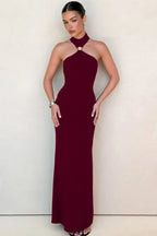 Woman wearing a burgundy sleeveless halter maxi dress with a gold-tone hardware detail at the neckline, standing against a neutral wall.