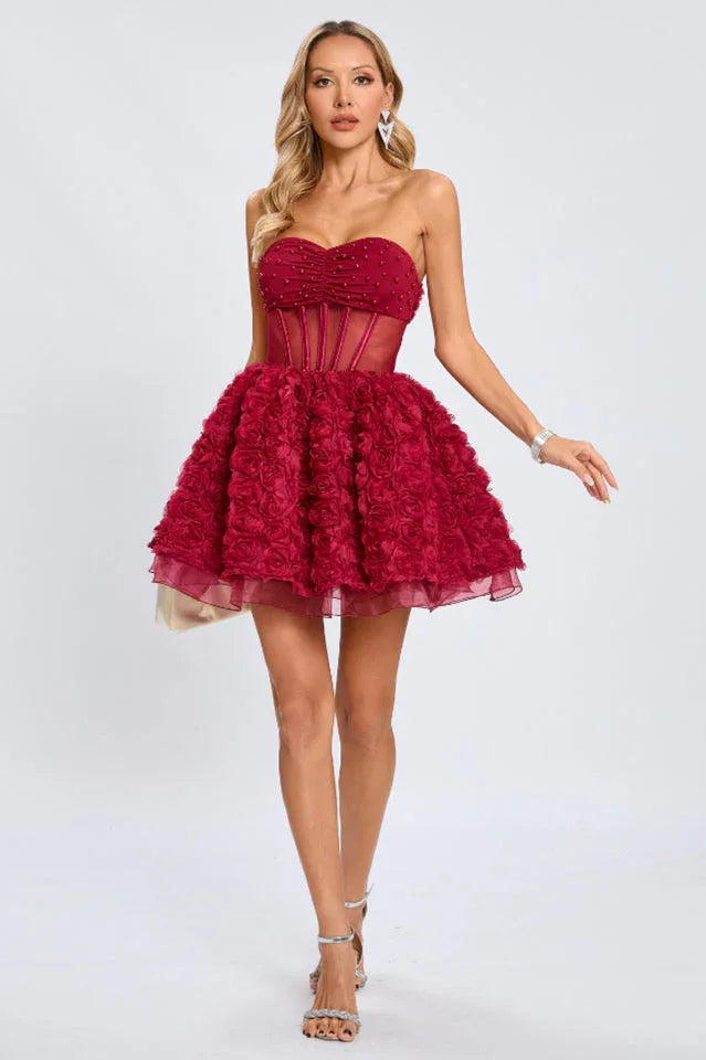 A woman in a strapless red dress with a voluminous floral skirt and sheer overlay, standing against a white background.