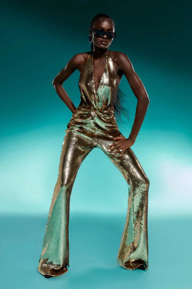 Model wearing a gold sequined halter neck jumpsuit with wide leg pants against a teal background.