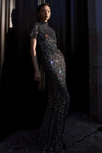 Model in a black sequined gown with a high neckline and short sleeves, standing in a dimly lit area.