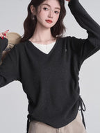 Woman in a dark grey v-neck sweater and tan pants, adjusting the drawstring on the side of her sweater.