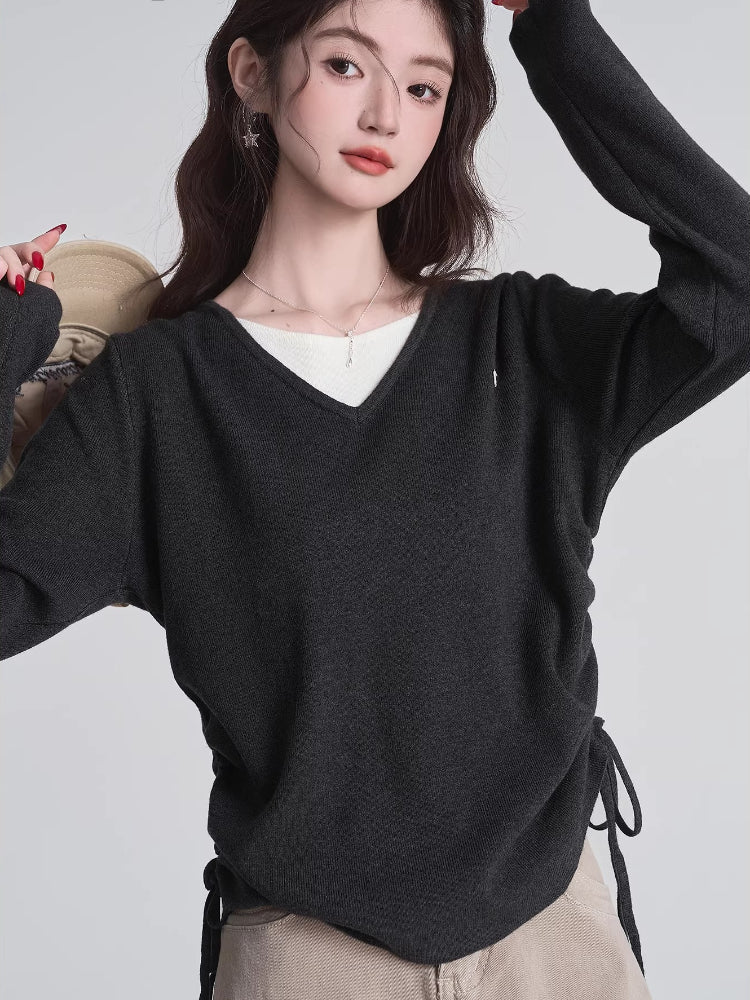 Woman in a dark grey v-neck sweater and tan pants, adjusting the drawstring on the side of her sweater.