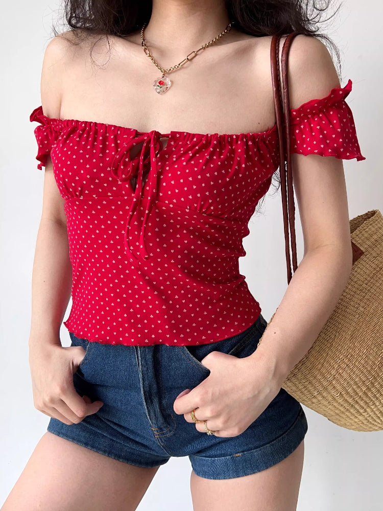 Woman posing with hands in pockets, wearing a red off-the-shoulder top and denim shorts