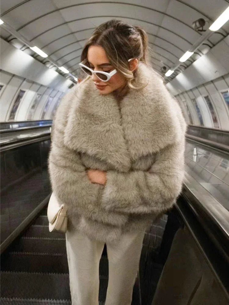 Woman wearing a light grey oversized faux fur jacket with white pants and white sunglasses on an escalator