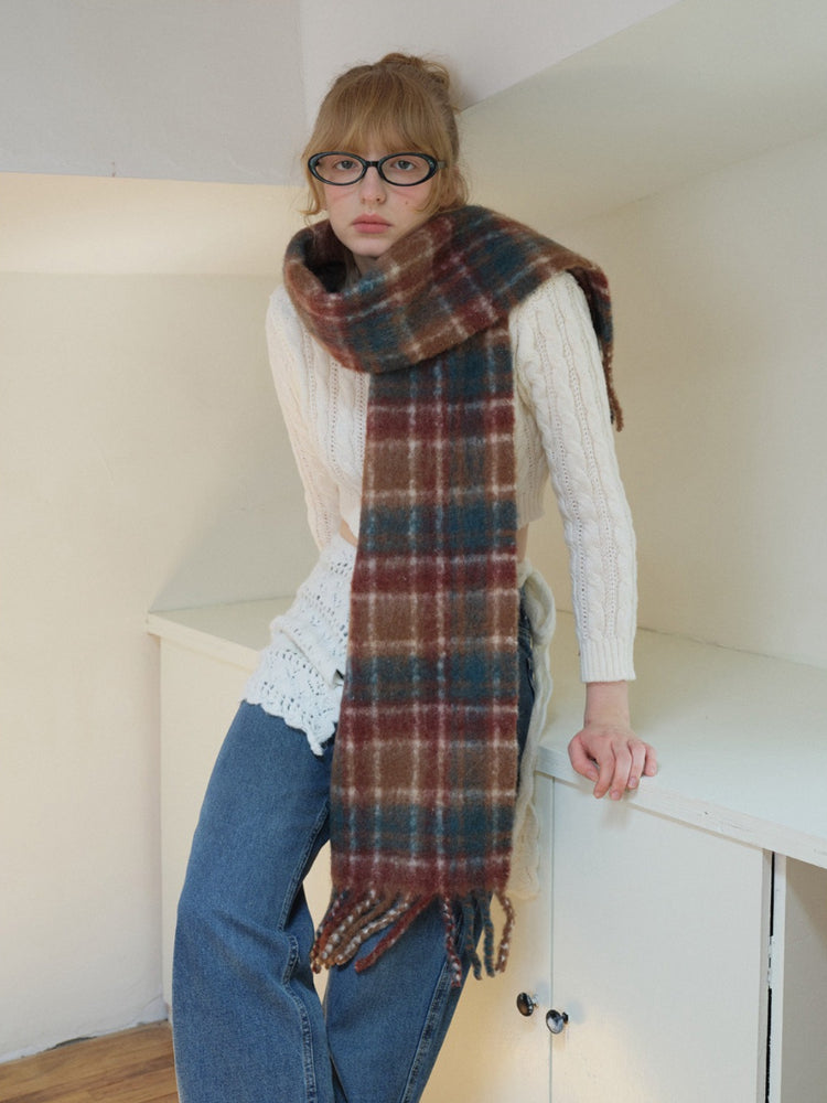 Close-up of a model wearing a white cable knit cropped sweater, blue denim jeans, a plaid scarf, and glasses, leaning against a shelf.