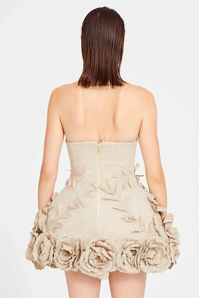 Back view of a woman wearing a strapless beige dress with a zipper closure. The skirt features a dense arrangement of fabric flowers around the hem.