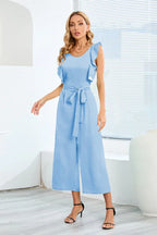Woman posing in a light blue wide leg culotte jumpsuit with ruffle sleeves and a self-tie waist, paired with black strappy heels.