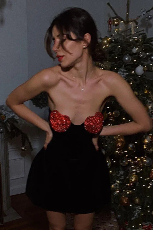 Woman in a black velvet strapless mini dress with red heart bust details, posing in front of a Christmas tree.