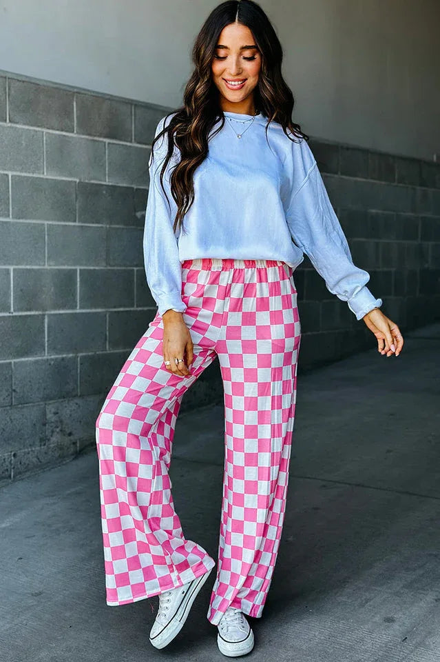 Woman posing in pink and white checkerboard wide leg pants with a light blue long-sleeved top and white sneakers.