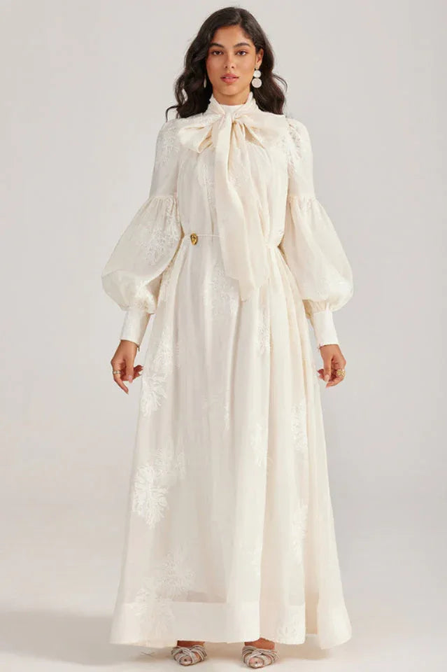 Woman wearing a long, off-white dress with a pussy-bow neckline and bishop sleeves, featuring an embroidered floral pattern.