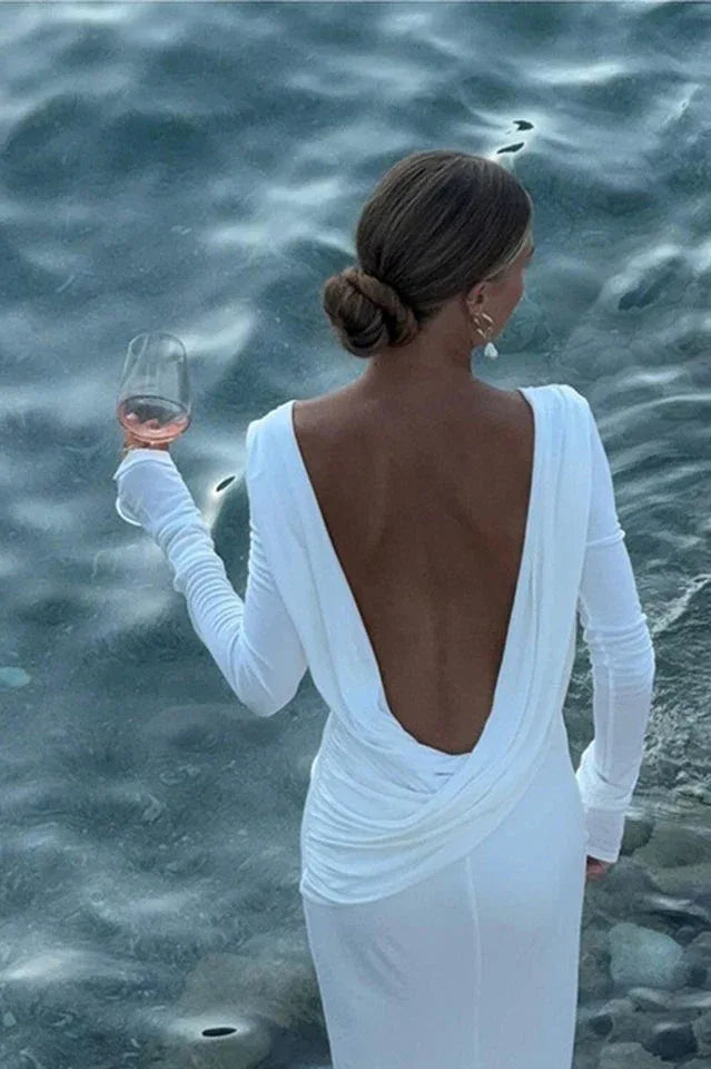 Woman in a white long-sleeved dress with a deep V-back, holding a wine glass by the water.
