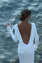 Woman in a white long-sleeved dress with a deep V-back, holding a wine glass by the water.