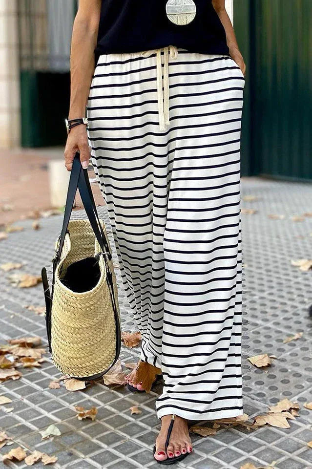 Close-up of a woman's legs wearing black and white striped wide leg pants and sandals, holding a straw tote bag.