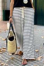 Close-up of a woman's legs wearing black and white striped wide leg pants and sandals, holding a straw tote bag.