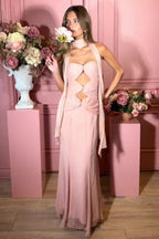 Woman wearing a pale pink halter neck maxi dress with cut-out details and a mermaid hemline, holding a flower in a pink studio setting.