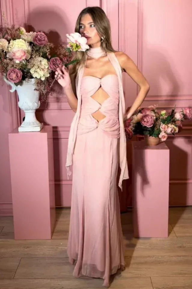 Woman wearing a pale pink halter neck maxi dress with cut-out details and a mermaid hemline, holding a flower in a pink studio setting.