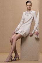 Full-length shot of a model in a cream-colored, long-sleeved shift dress adorned with crystals and pearls, paired with silver strappy heels.