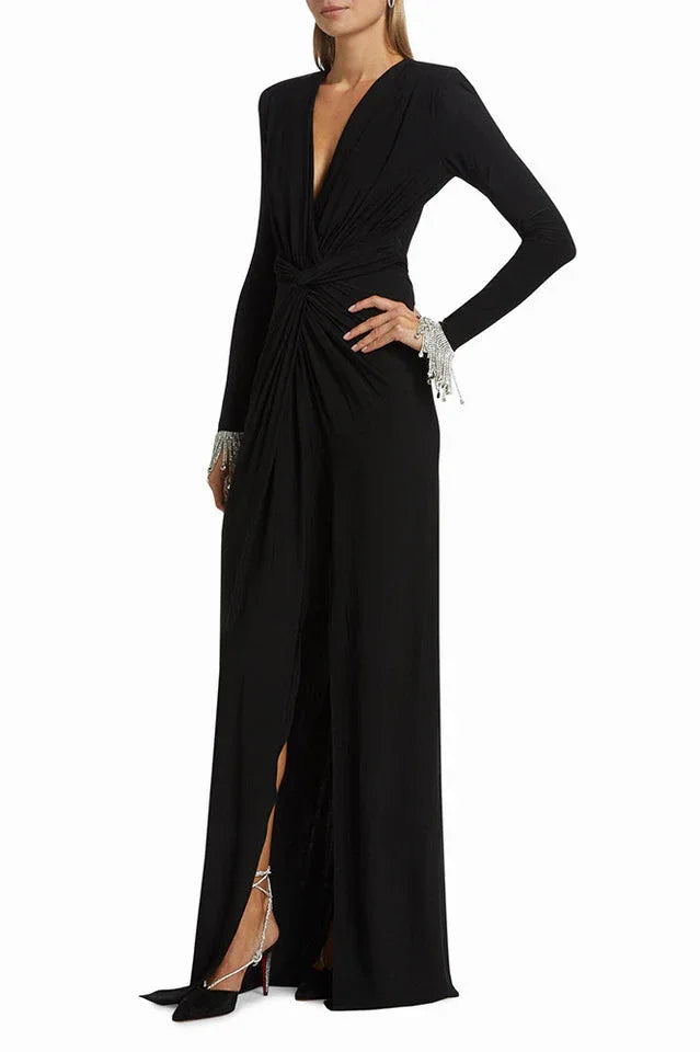 Woman posing in a long-sleeved black gown with a plunging V-neckline, ruched bodice, thigh-high slit, and embellished cuffs, showing the side profile.