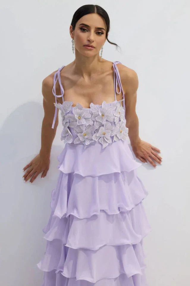 Woman leaning against a white wall, wearing a light purple tiered ruffled chiffon dress with a floral applique bodice and tie shoulder straps.