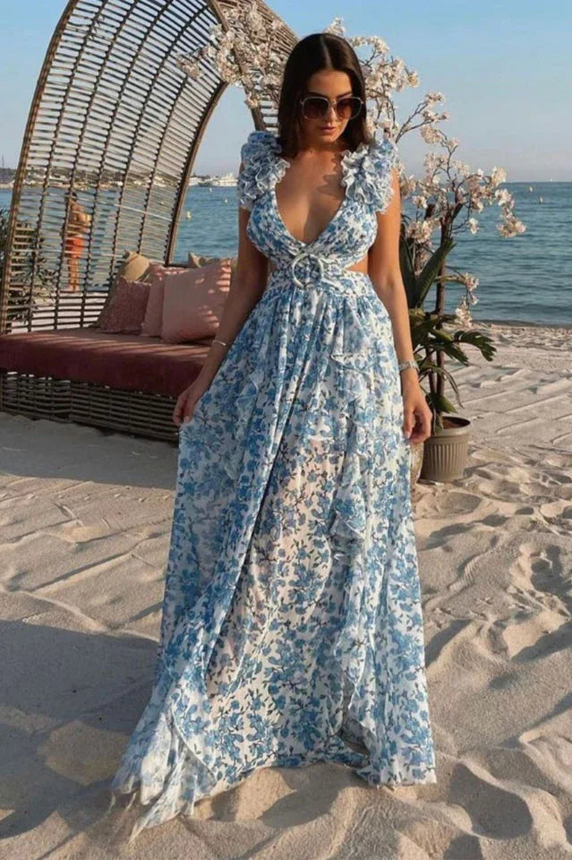 Woman wearing a blue and white floral print maxi dress with ruffled sleeves and cut-out details, standing on a sandy beach.