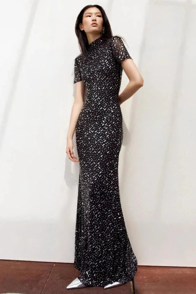 Woman in a black sequined gown with a high neckline and short sleeves, posing against a white wall.